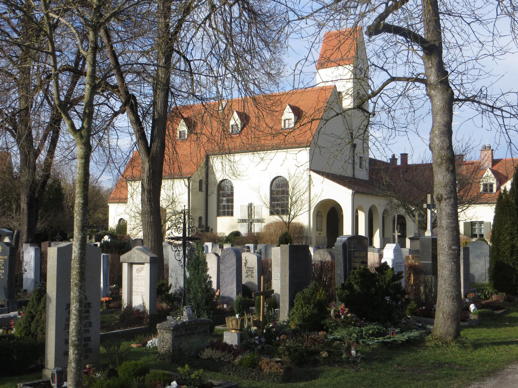 Grabreihen zwischen hohen Bäumen vor einer weißen Kapelle mit rotem Dach auf Friedhof Pasing