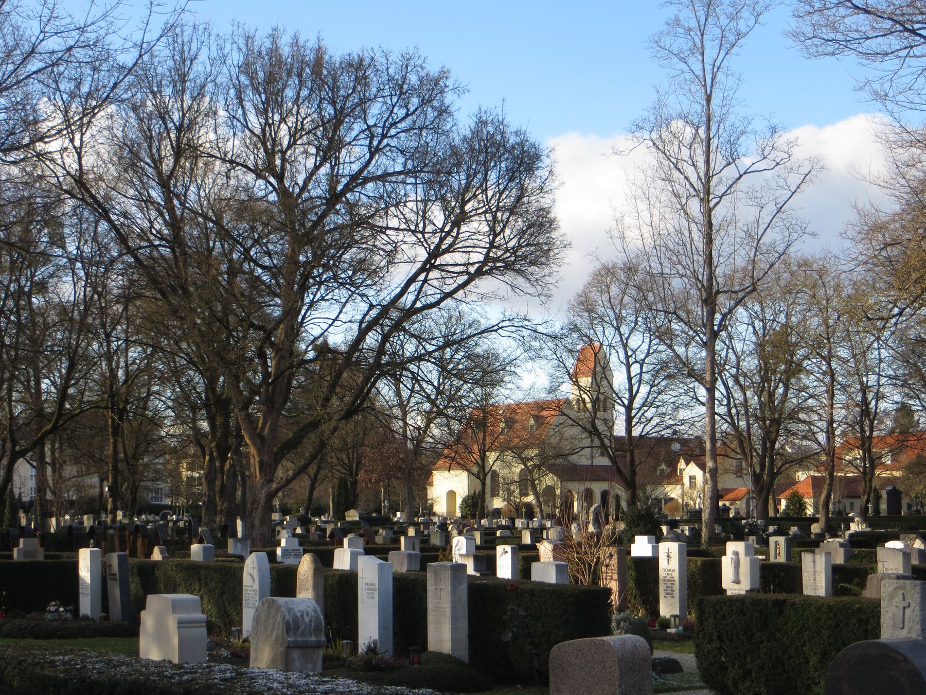 Weiter Blick über Grabreihen unter laublosen Bäumen zur weißen Kapelle auf Friedhof Pasing