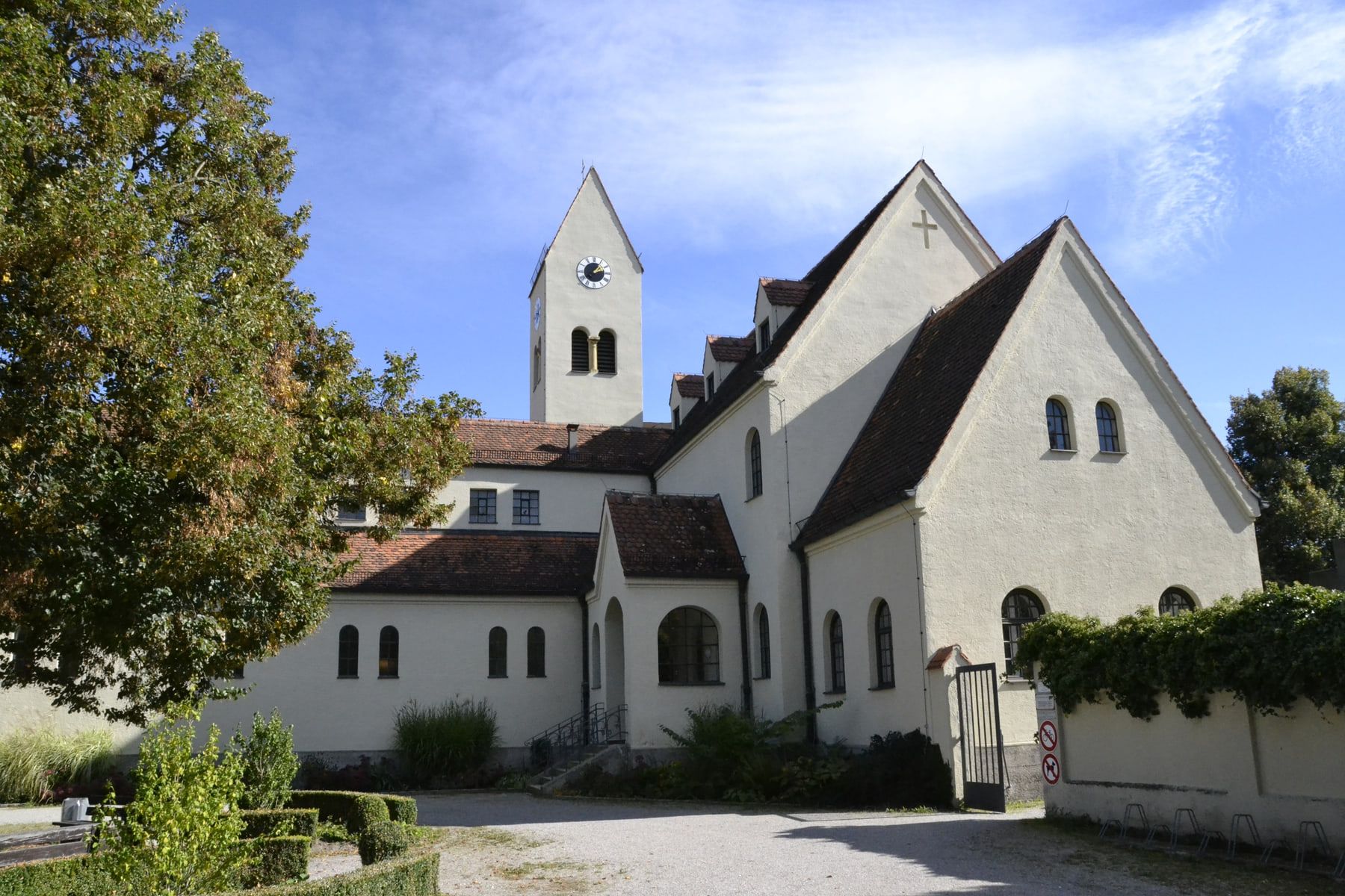 Weißer Gebäudekomplex mit steilen dunklen Dächern, Turm und gepflastertem Vorplatz auf Friedhof Pasing